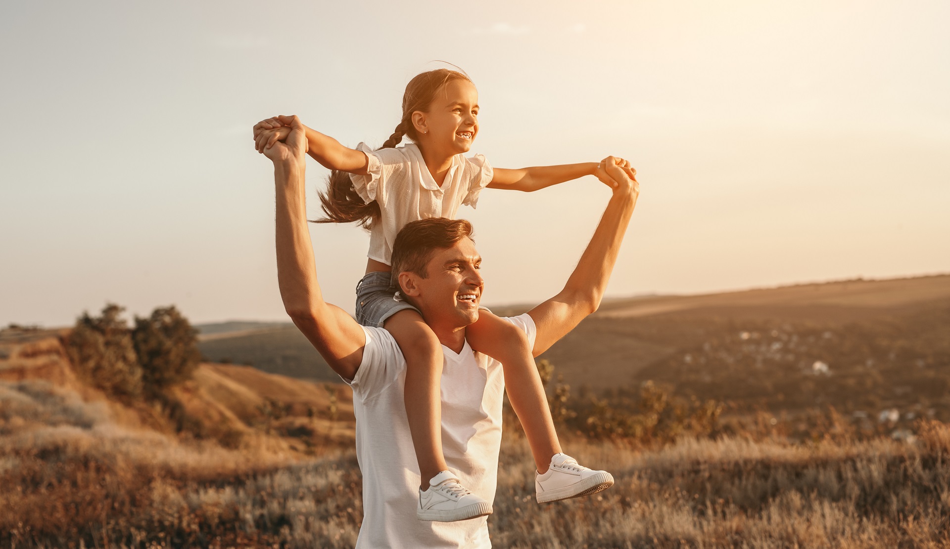 A imagem mostra um homem com uma criança em seu pescoço, e estão sorridentes. O homem vesti uma blusa branca e a criança short azul, tênis branco, blusa rosa. Ao fundo da imagem tem montanhas e vegetação baixa, e está ensolarada.